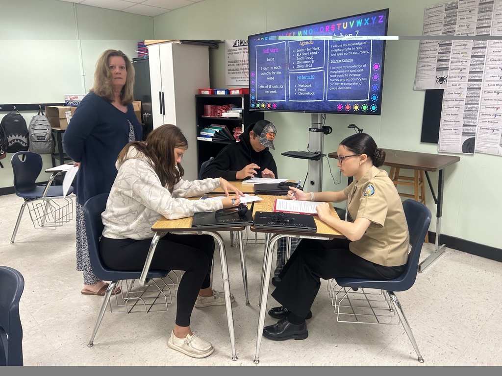 Three people, one standing and two sitting, at a desk with a monitor in a classroom.