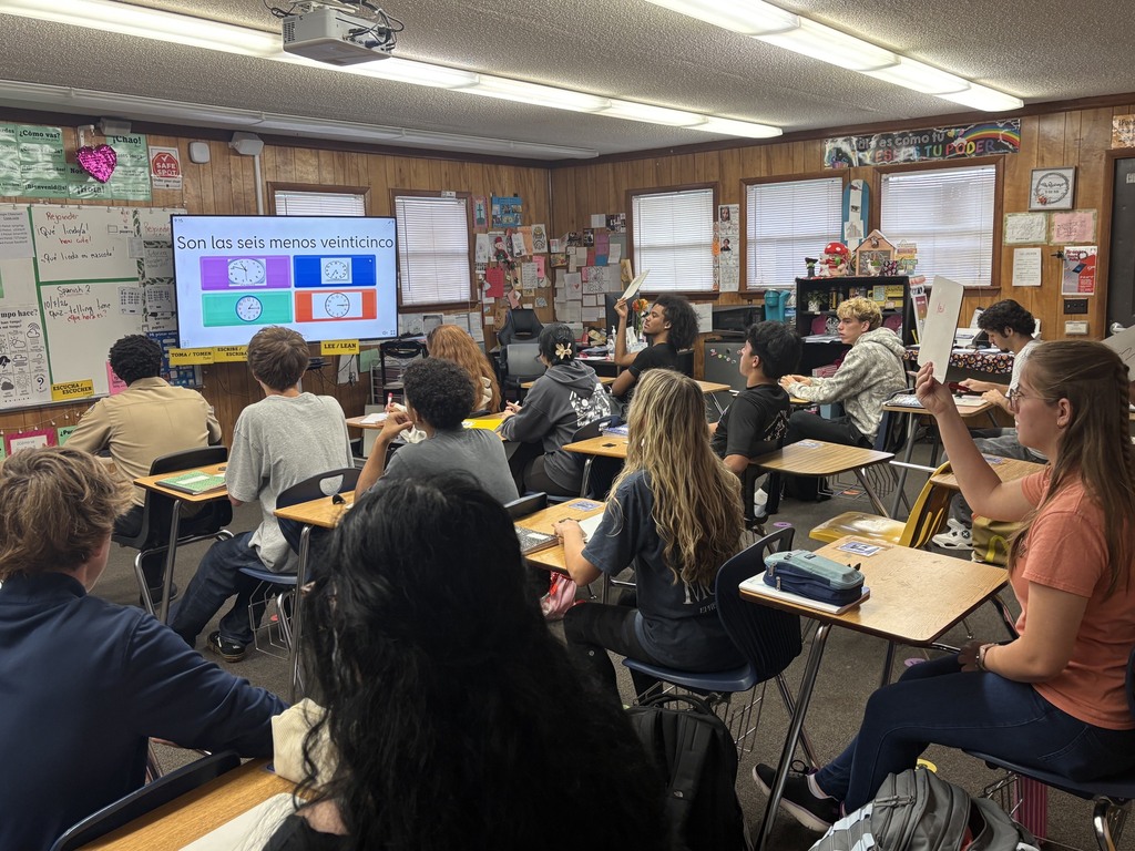 A classroom with students seated around desks facing a front screen. A teacher leads a discussion.