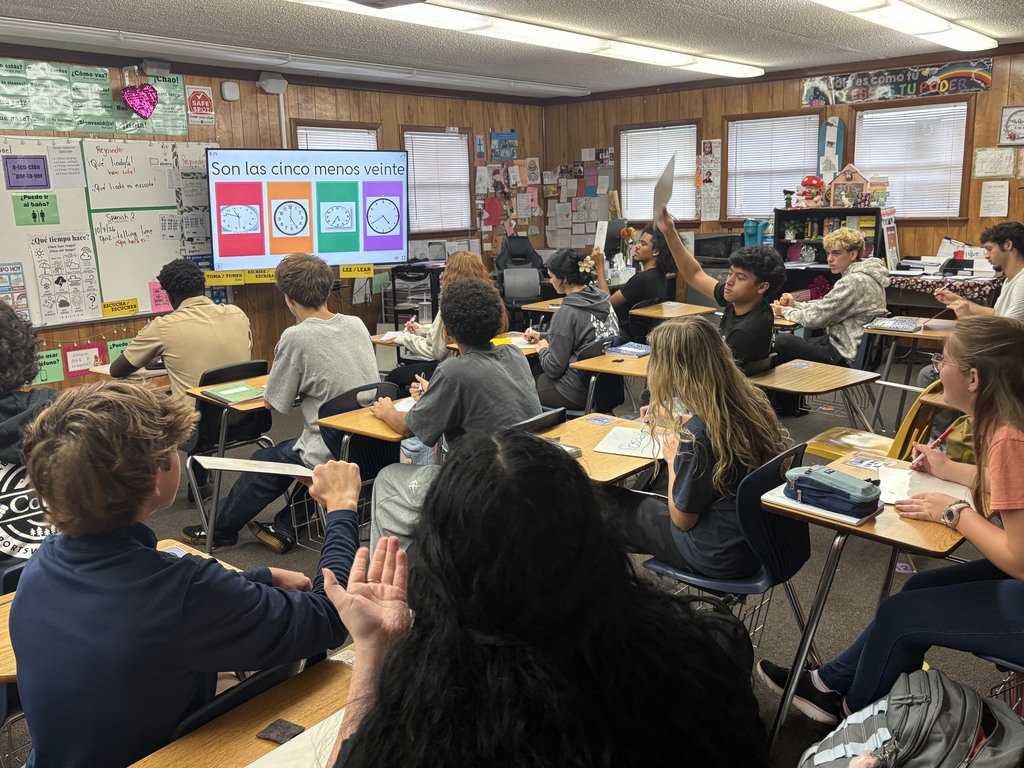 Students in a classroom with wooden walls, desks, and a projector screen. Some students sit with hands raised.