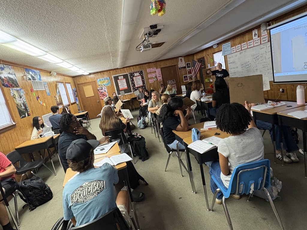 A classroom with students seated in rows at desks. A teacher stands near a projector screen and whiteboard.