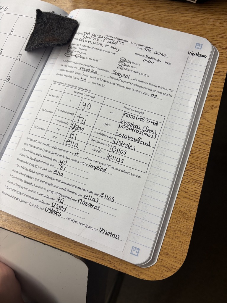 An open notebook with lined pages and handwritten notes sits on a wooden table. A small piece of dark paper is placed on the left side of the notebook.