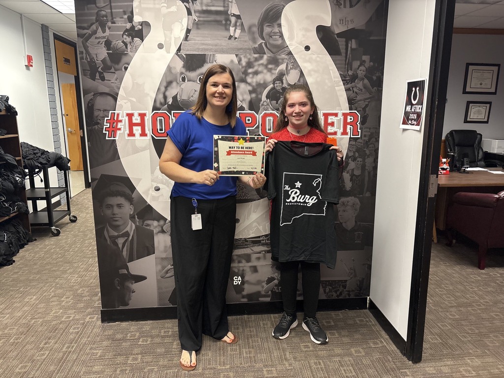 Two women stand before a large mural. One holds an award, the other a shirt. The background features a horseshoe and photos.