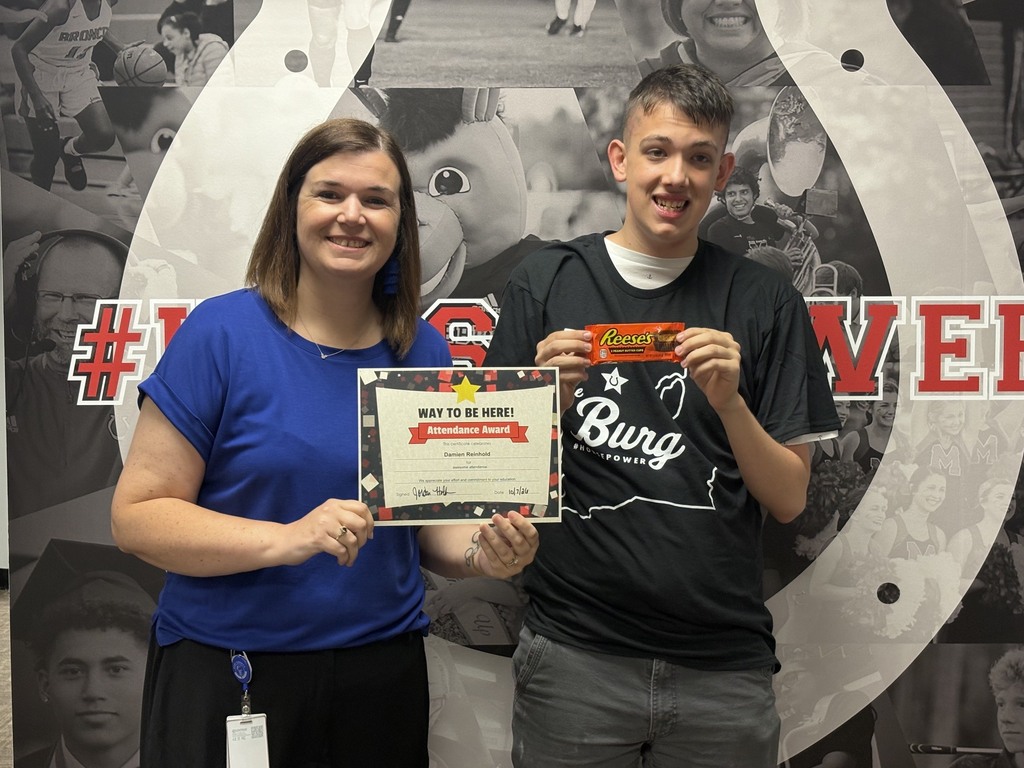 Two people, a woman and a man, stand smiling with a certificate. Behind them, a large horseshoe with team logos and images.
