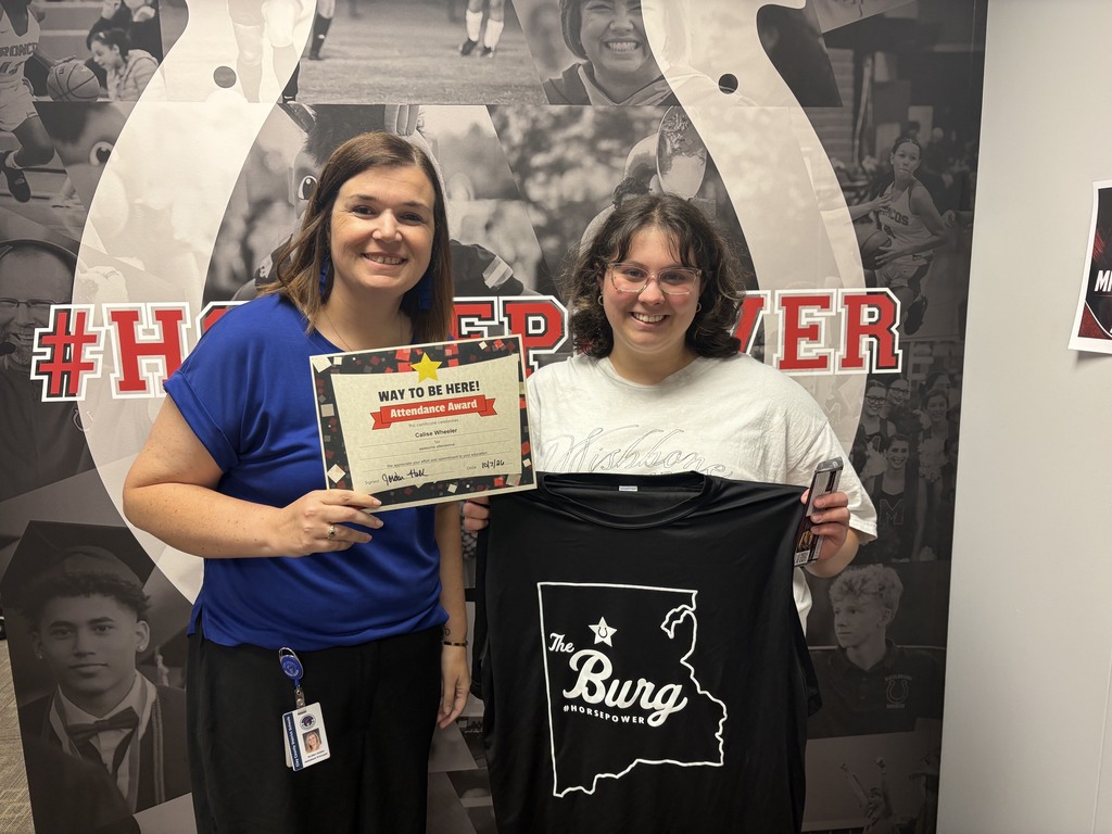 Two women pose with a certificate and a black shirt against a wall with a horseshoe and black and white photos.