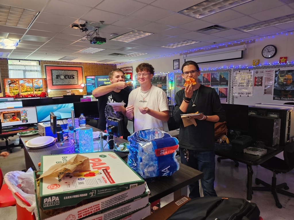 Three people in a room with monitors and tables, each holding a slice of pizza.