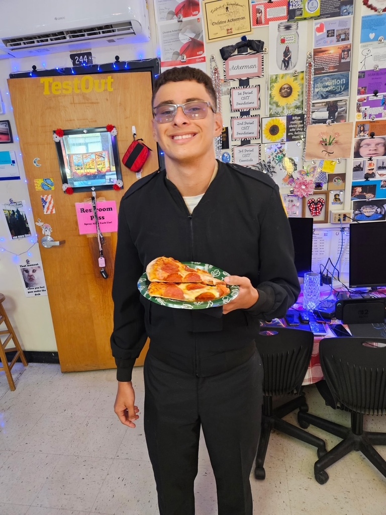 Man with glasses in a black uniform holds a plate of pizza in a room with chairs, monitors, and bulletin board.