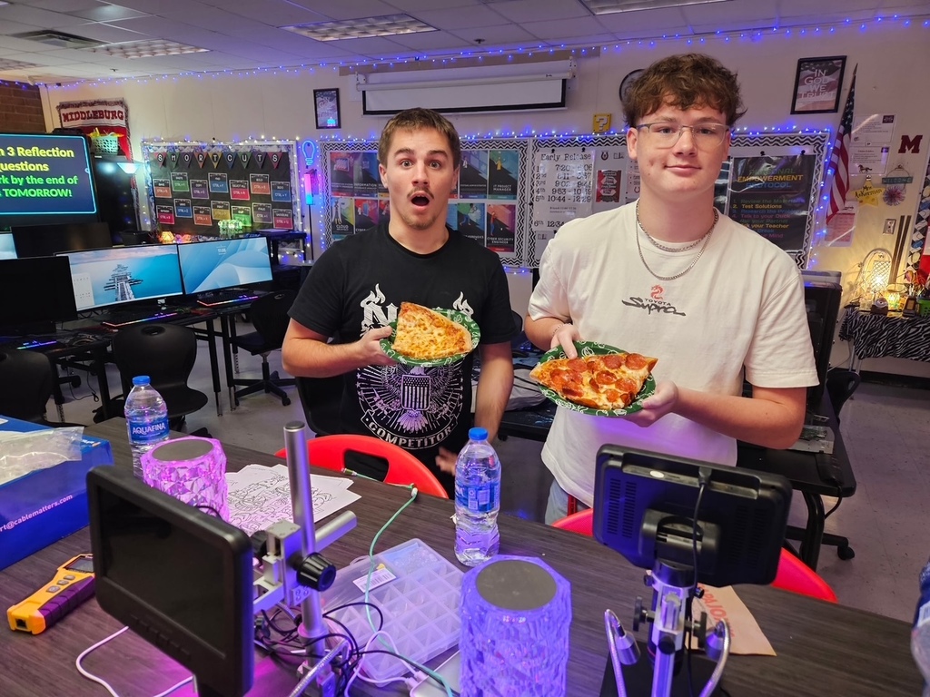Two people in a gaming room pose with pizza. One holds pizza, and the other opens mouth wide.