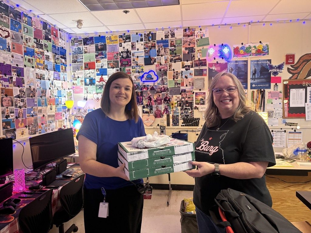 Two women hold boxes in a room with colorful posters on walls, computers, and a desk.