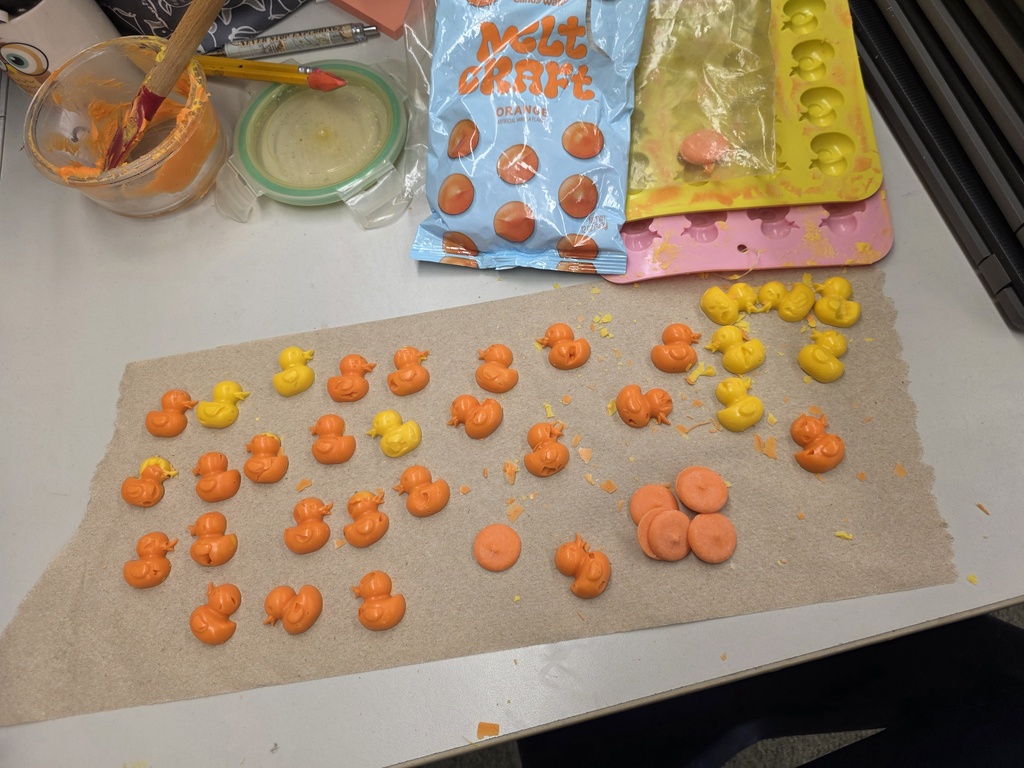 A table displays multiple small orange and yellow items on parchment paper. Paints, brushes, and molds are nearby.