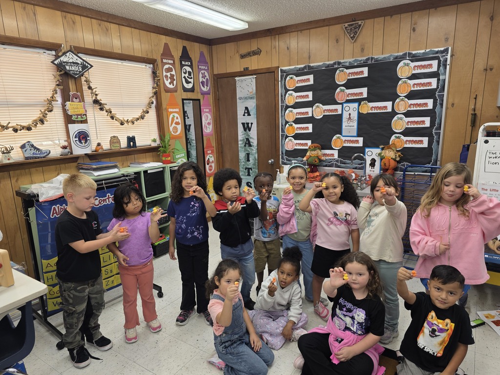 Kids in a classroom with bright walls and decorations, some standing and others seated, all smiling.