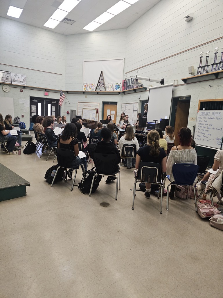 A classroom with students seated in chairs, some in rows, a whiteboard on the wall, and sunlight coming through windows.
