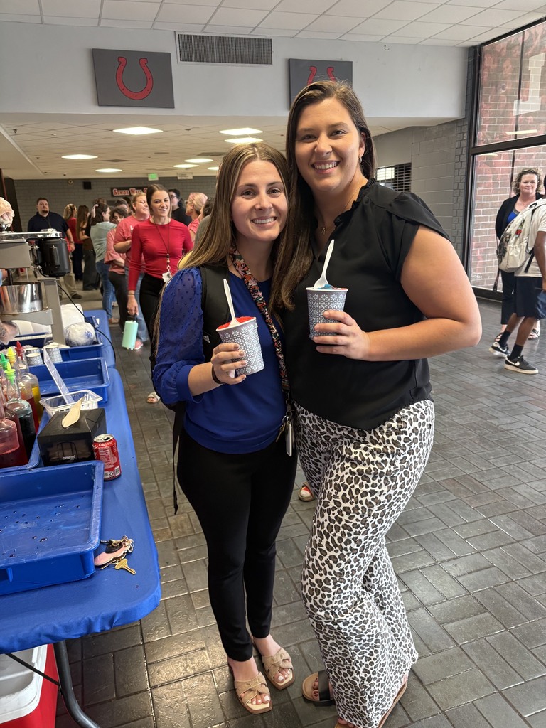 Two women, one in blue, the other in black with leopard print pants, stand smiling with drinks in a cafeteria.