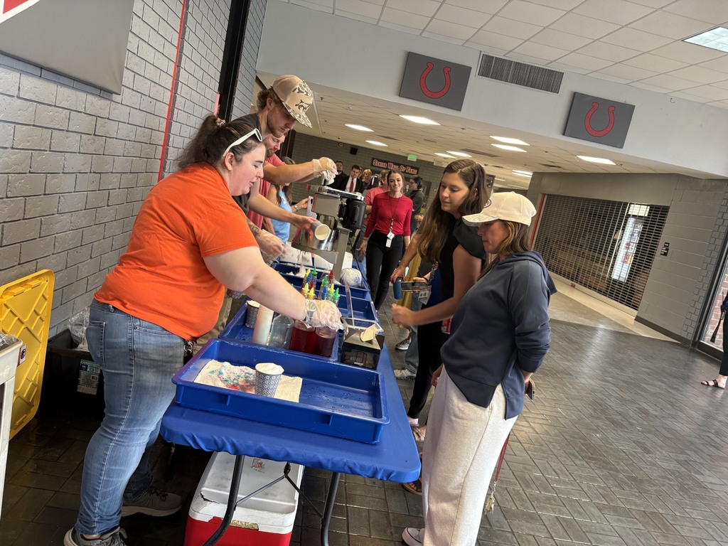 A group of people are preparing food at a table in a school hallway, with one person wearing a hat.