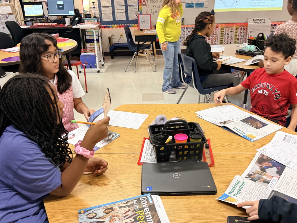 Several students seated at a table, engaged in reading newspapers and writing. Background contains tables, chairs, and a projector screen.