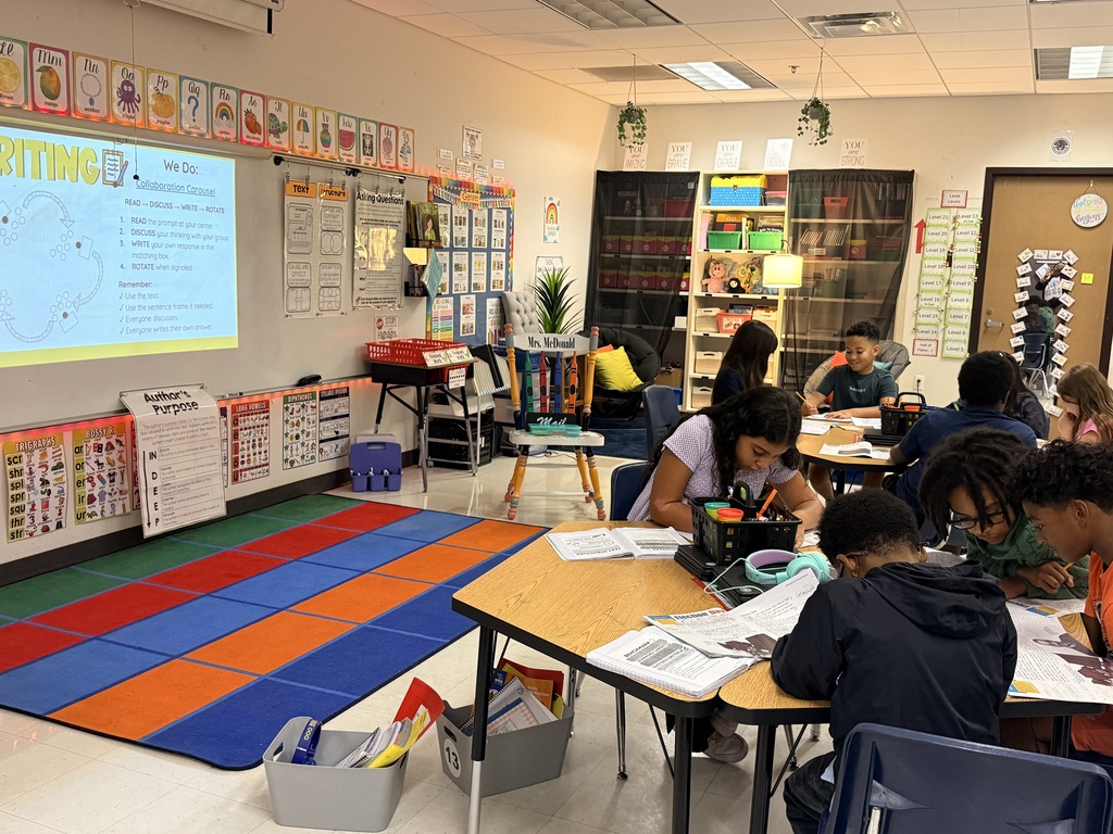 Classroom with students seated at tables, projector screen on wall, and colorful rug on floor.