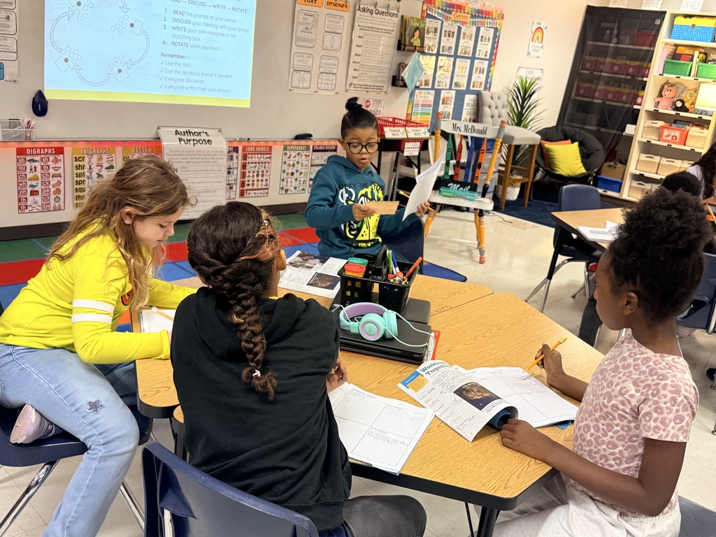 Four students sit around a table, reading and writing in a classroom. One wears a yellow sweatshirt.