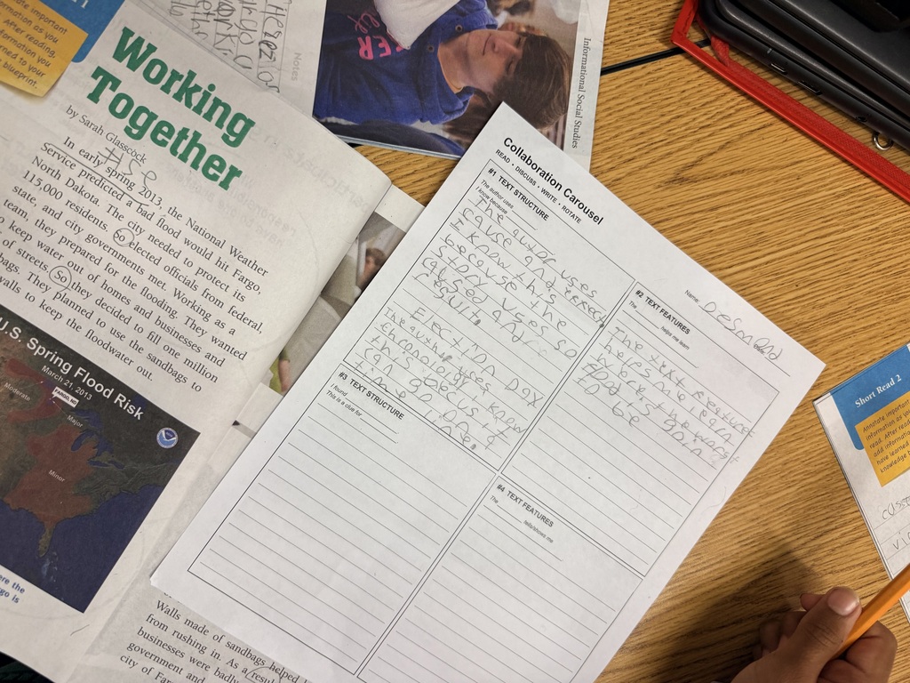 A person holds a paper with lined text and an open newspaper with a blue-clad figure on a wooden desk.