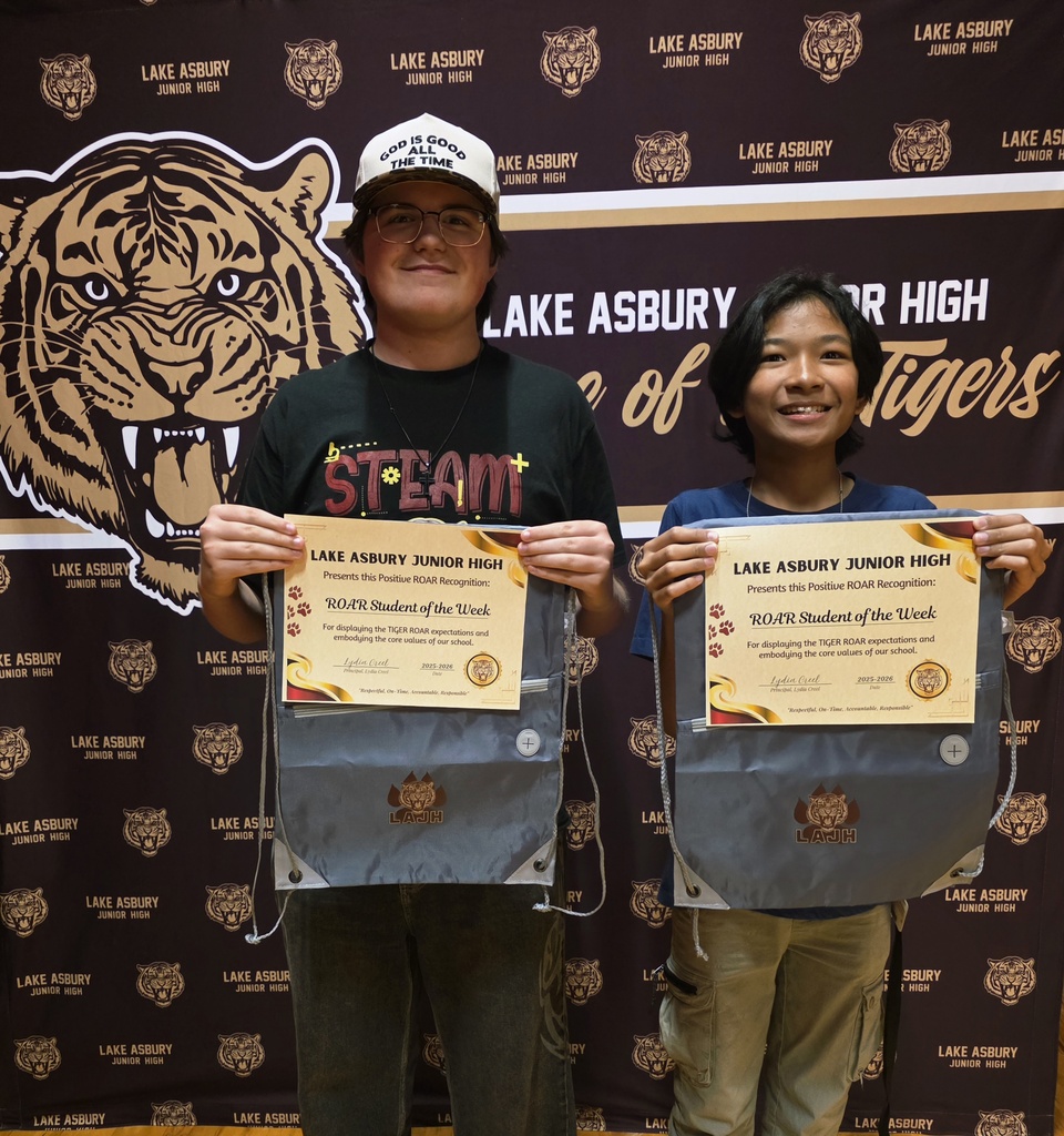 Two students, one wearing glasses and a cap, hold up certificates with school logos. They stand before a wall with a tiger logo.