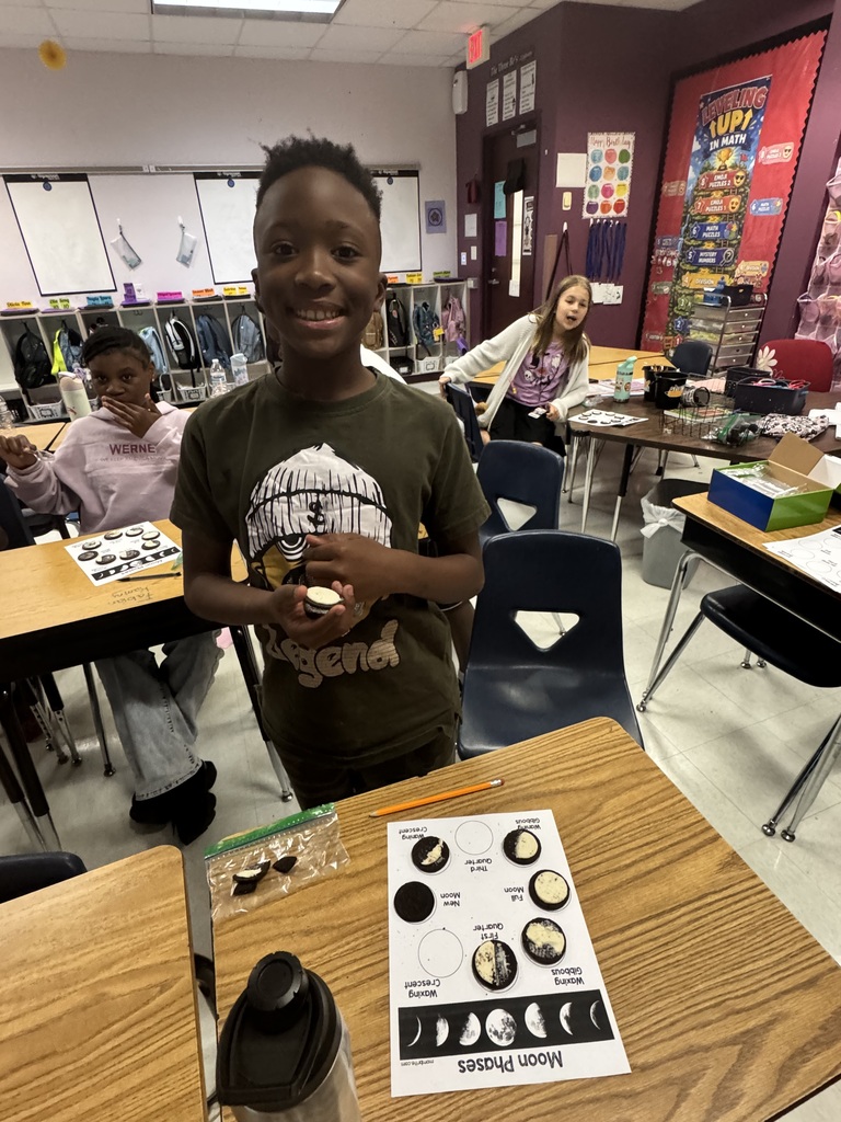 A student in a green T-shirt stands in a classroom, smiling. He holds a small object and stands by a desk with a black and white chart.