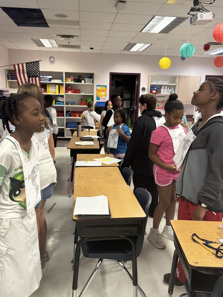 Students in lab coats gather in a classroom with desks, tables, and an American flag.