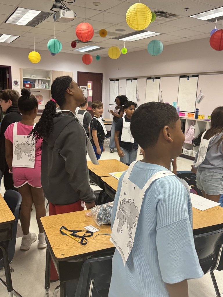 A classroom with colorful paper decorations. Students wear aprons with white papers on their backs.