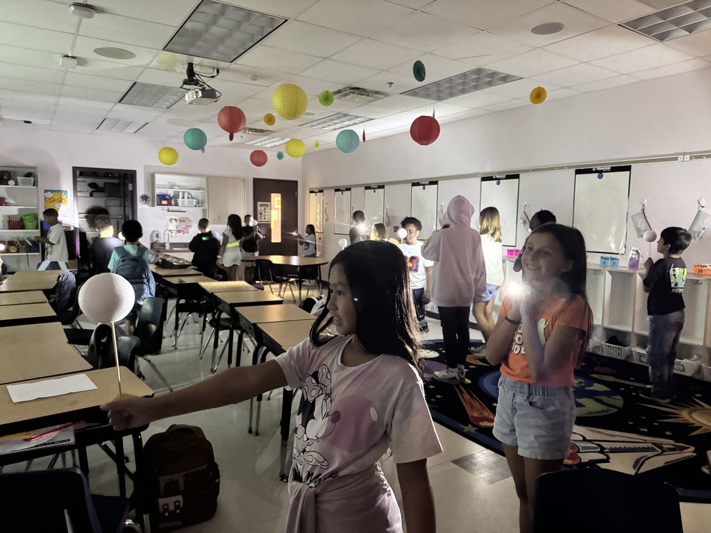 Classroom with students. A girl stretches out her arm to hold a balloon. Balloons float from ceiling.