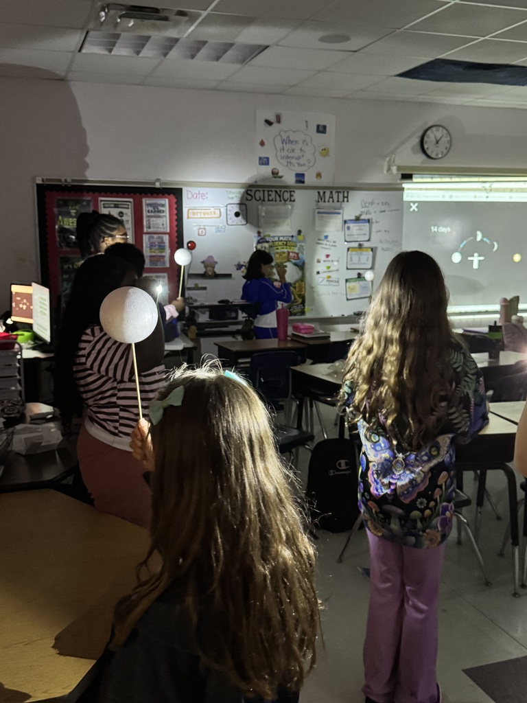 A classroom with a group of students. One holds a flashlight and another has long hair.