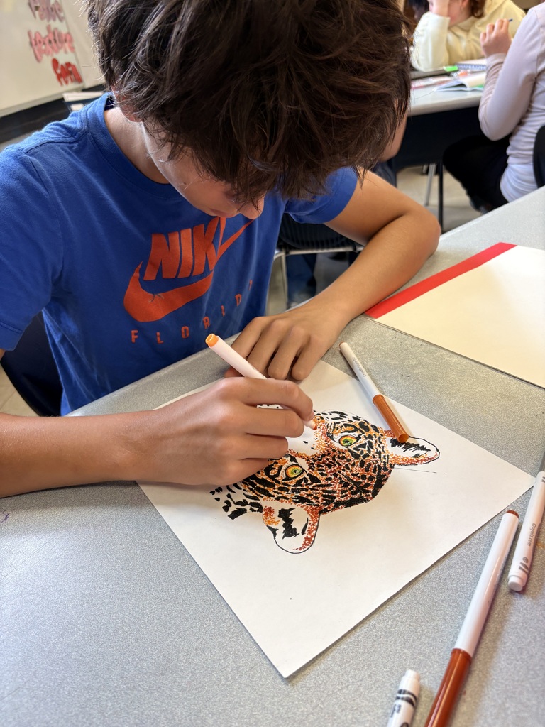 A boy in a blue shirt draws a tiger on paper using colored markers. Other people work at desks behind him.