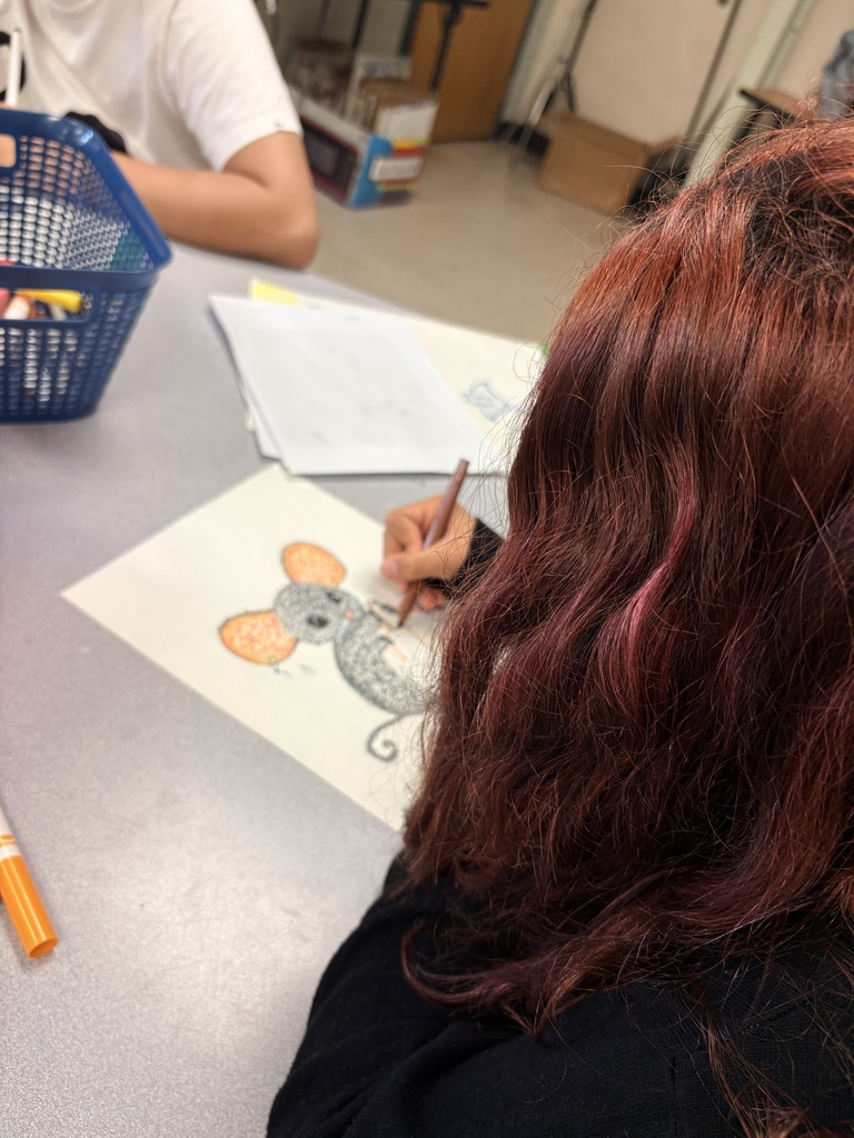 A person with long hair draws a mouse on a paper. Another person sits at the table behind.
