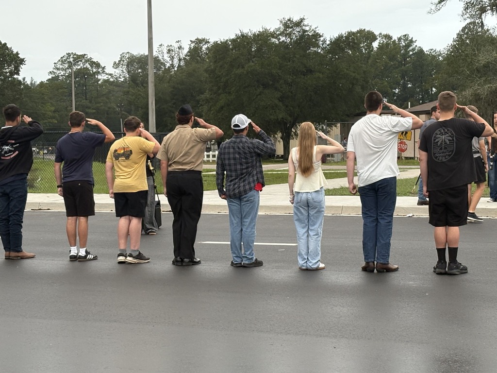 Several adults and teenagers stand in a line and salute. They are in a parking lot, with trees and a building in the background.