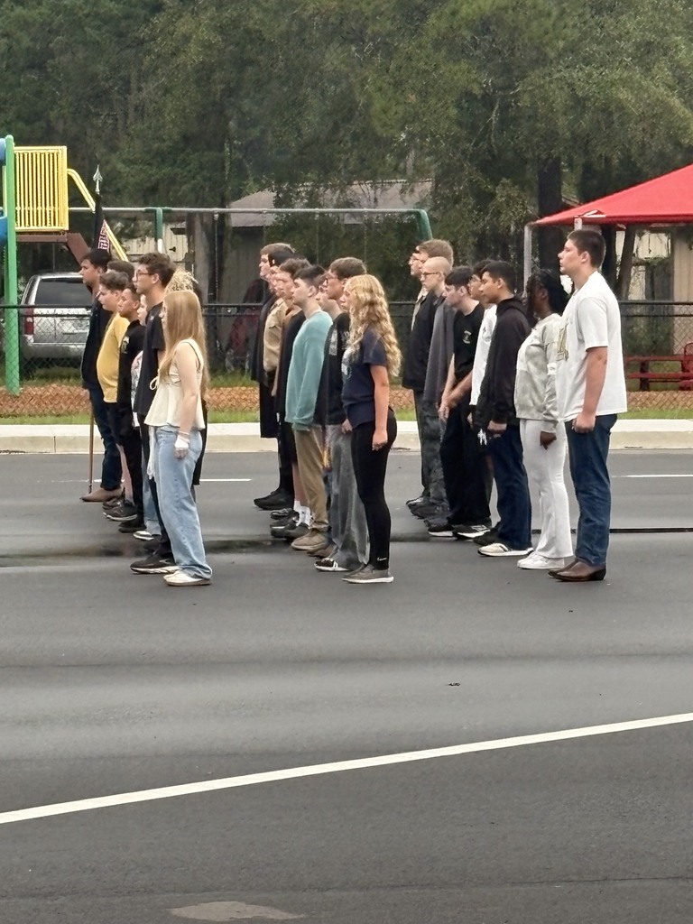 A group of people standing in line on a street. Behind them is a playground with a red canopy.