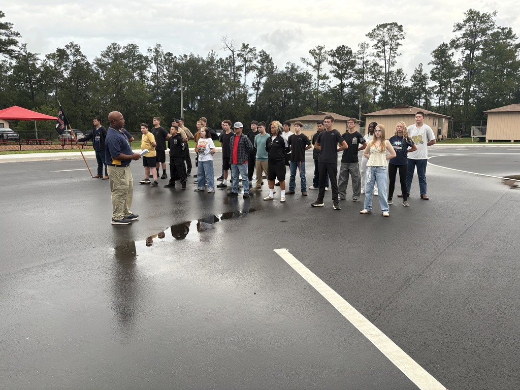 A group of people stands in a wet parking lot, with one person speaking in the center.
