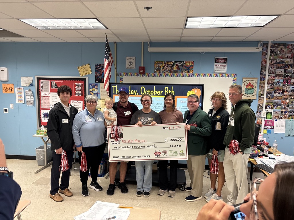 Several adults and children stand behind a large check. Some hold ribbons. A flag hangs behind them.