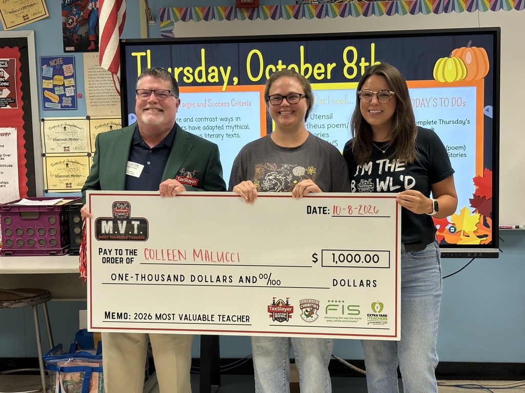 Three adults standing in a classroom holding a check for $1,000,00. Behind them is a whiteboard with the date and a pumpkin decoration.