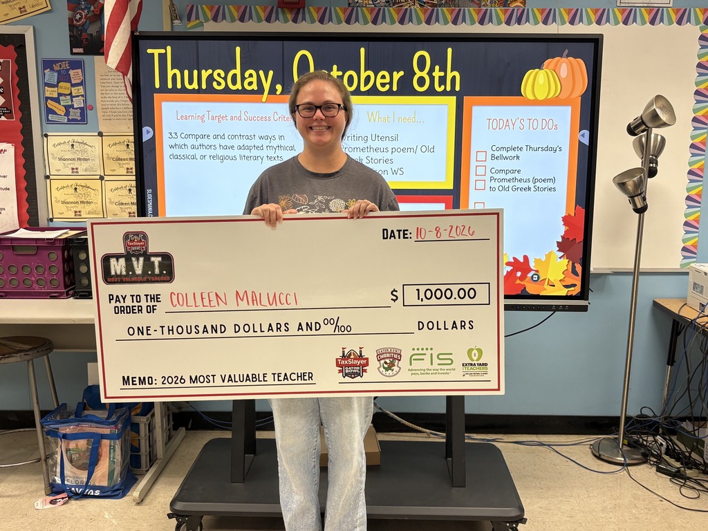 A person in glasses and a gray shirt holds a check for $1,000.00 labeled "Most Valuable Teacher." The background includes a classroom with a monitor and table.