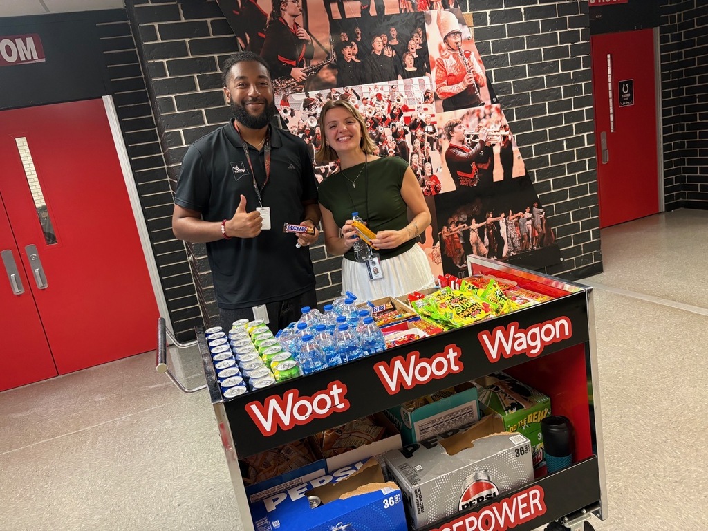 A man and woman next to a cart filled with snacks and drinks. Behind them is a brick wall with a collage.