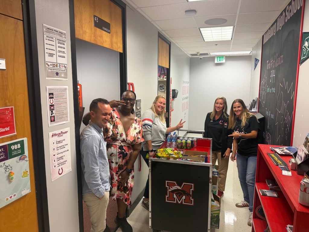 Six people in a hallway, with two women holding a bottle and others standing near a cart with the letter M.