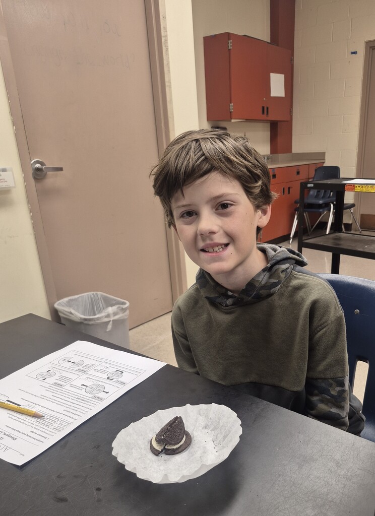 A boy smiles at a desk with an Oreo cookie in a paper holder. A pencil and papers are on the desk. A trash bin is beside the door.
