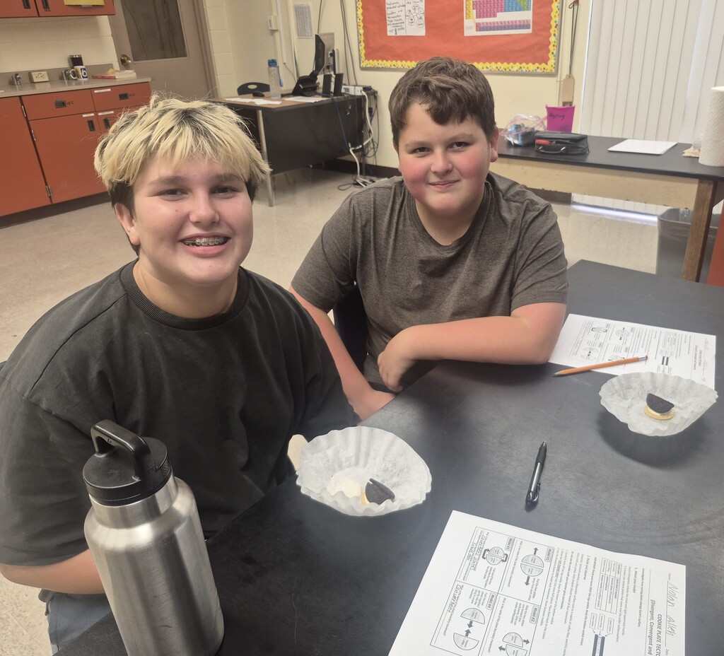 Two boys sitting at a table with papers and pencils in front of them. One boy has blonde hair and the other has brown hair.