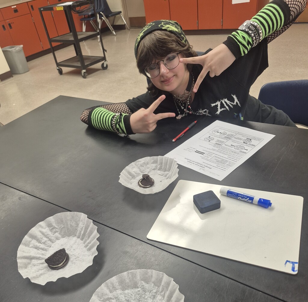 A person wearing glasses and a black shirt seated at a table, posing with a peace sign. Papers, coffee filters, and a pen are on the table.