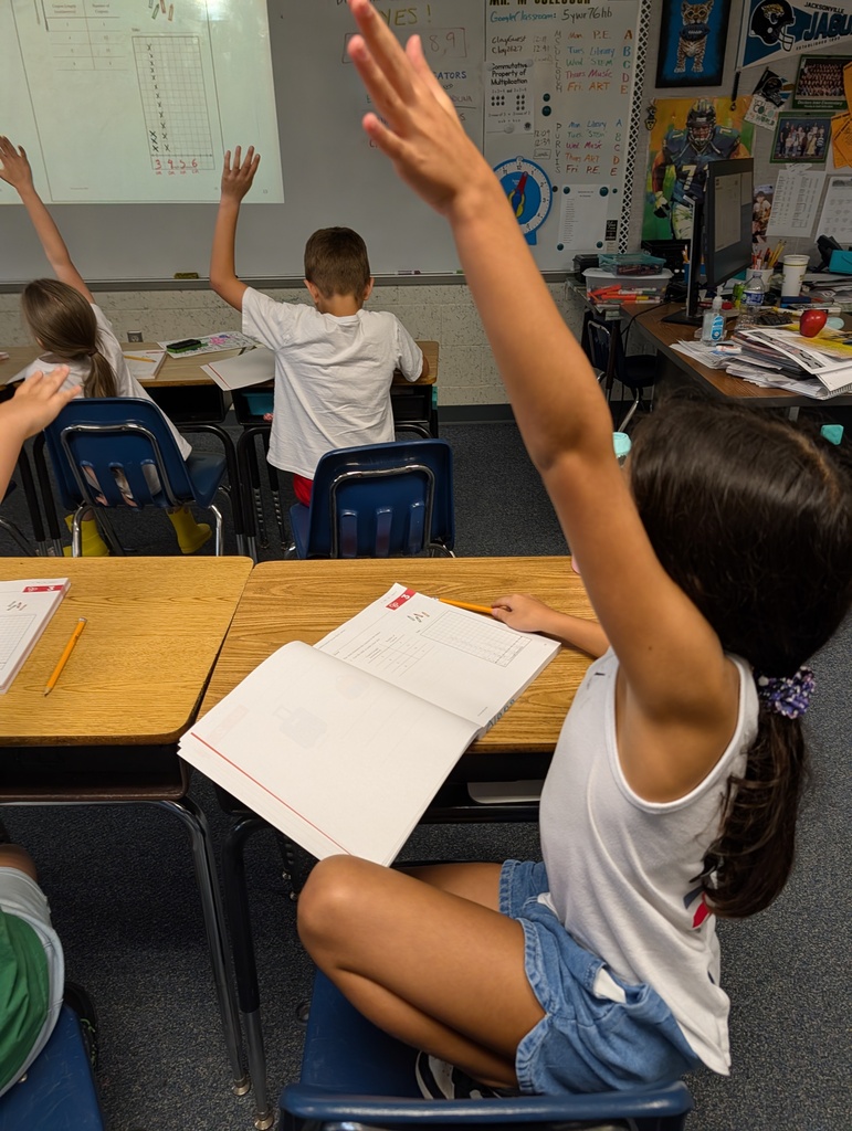 A classroom with children seated in chairs raising their hands. A projector screen displays a math problem.