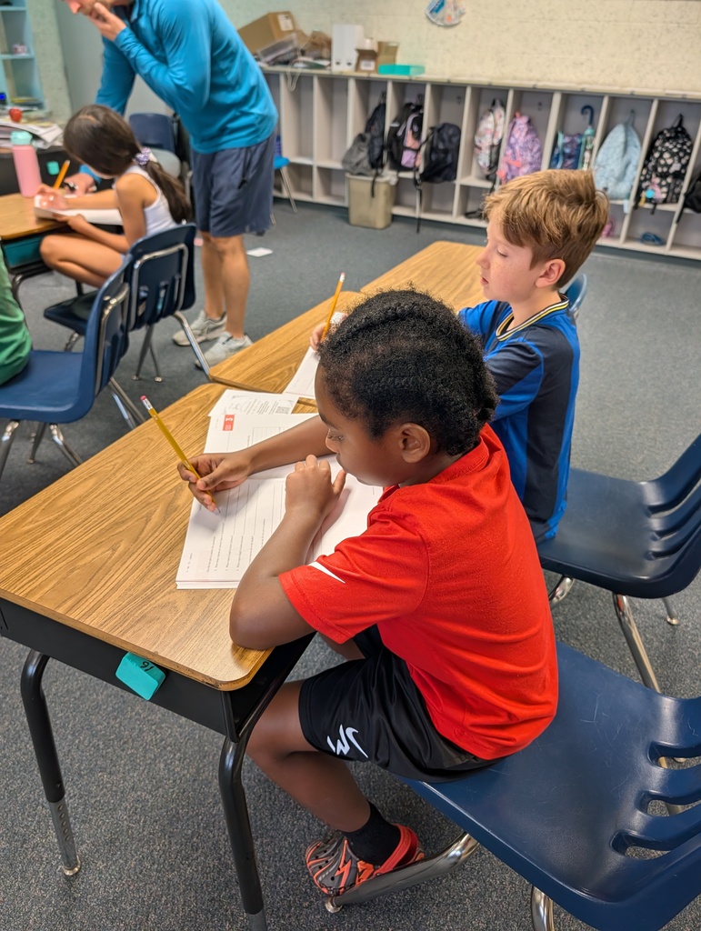 Two boys in a classroom, one wearing a red shirt, seated at a desk with papers.