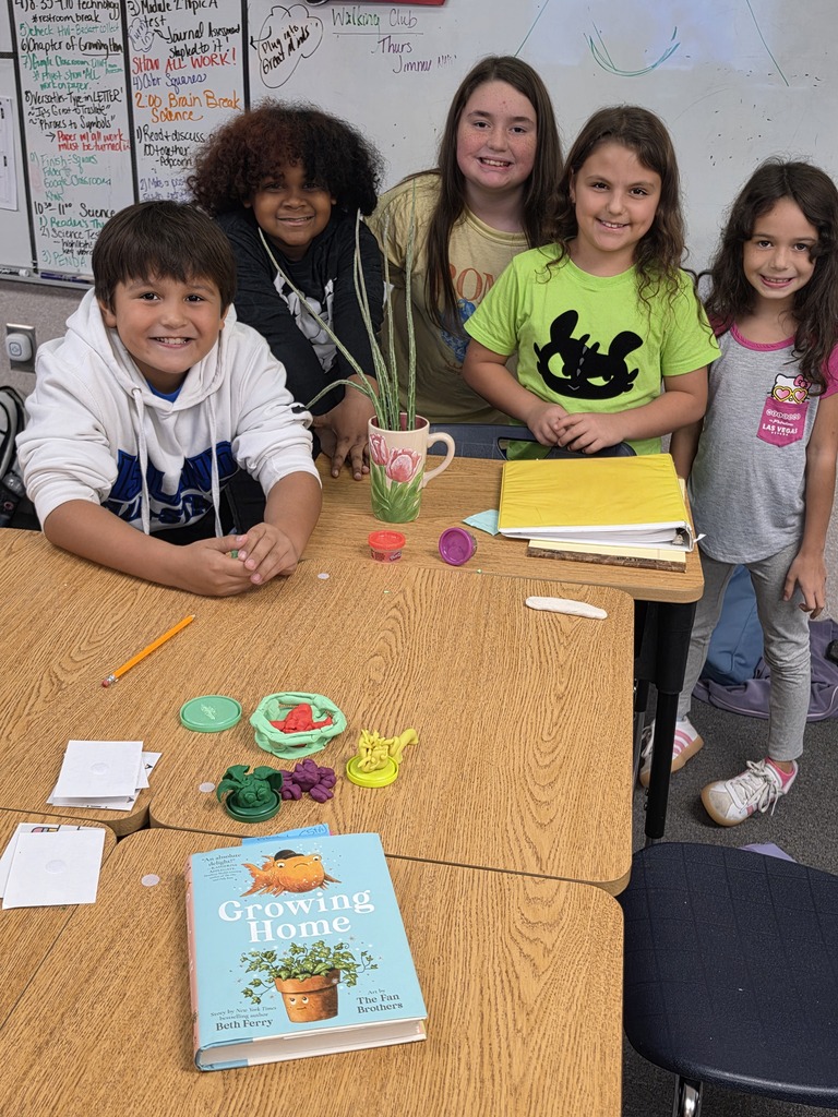 Five children, four standing and one sitting, are gathered around a table with books and small objects. The table is in a classroom with a whiteboard in the background.
