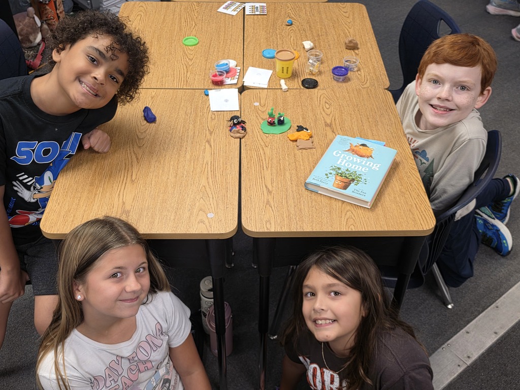 Four kids sitting at desks in a classroom, smiling and looking at the camera. Items on the tables include books, small toys, and a plant.