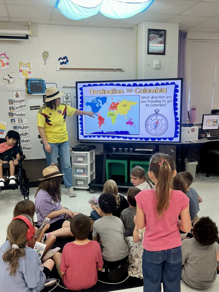 A teacher stands by a screen showing a map. Students sit on the floor, listening. The room has posters and books.