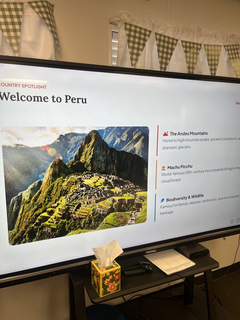 A large monitor displays an image of mountains. Text reads "Welcome to Peru" and "The Andes Mountains." Tissues are on the stand.