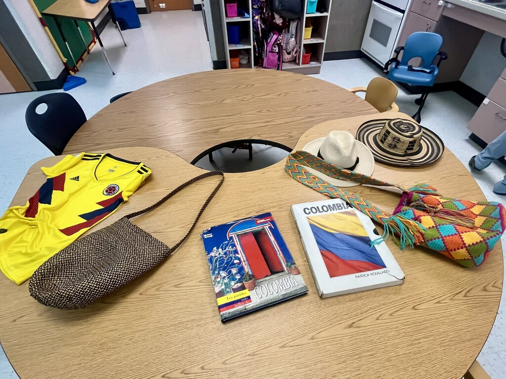 A round table displays various items: a colorful soccer jersey, a book, a hat, and two bags. Background includes a white wall and shelves with items.