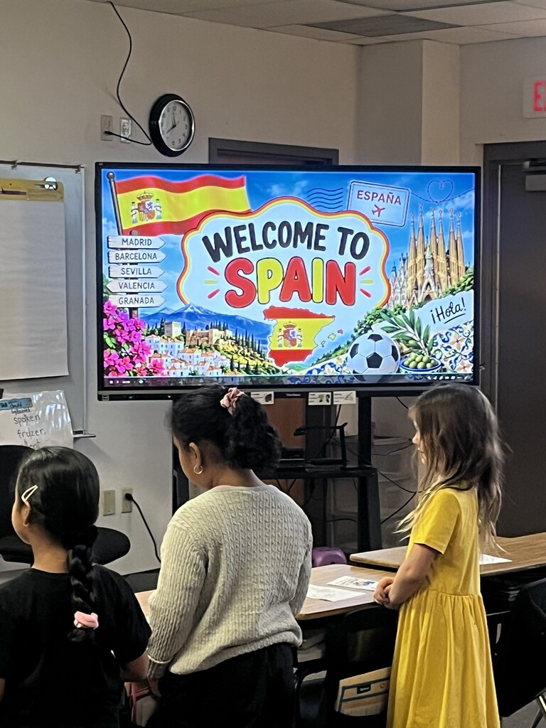 Three children sit at a desk in a classroom with a large monitor displaying a Welcome to Spain graphic.