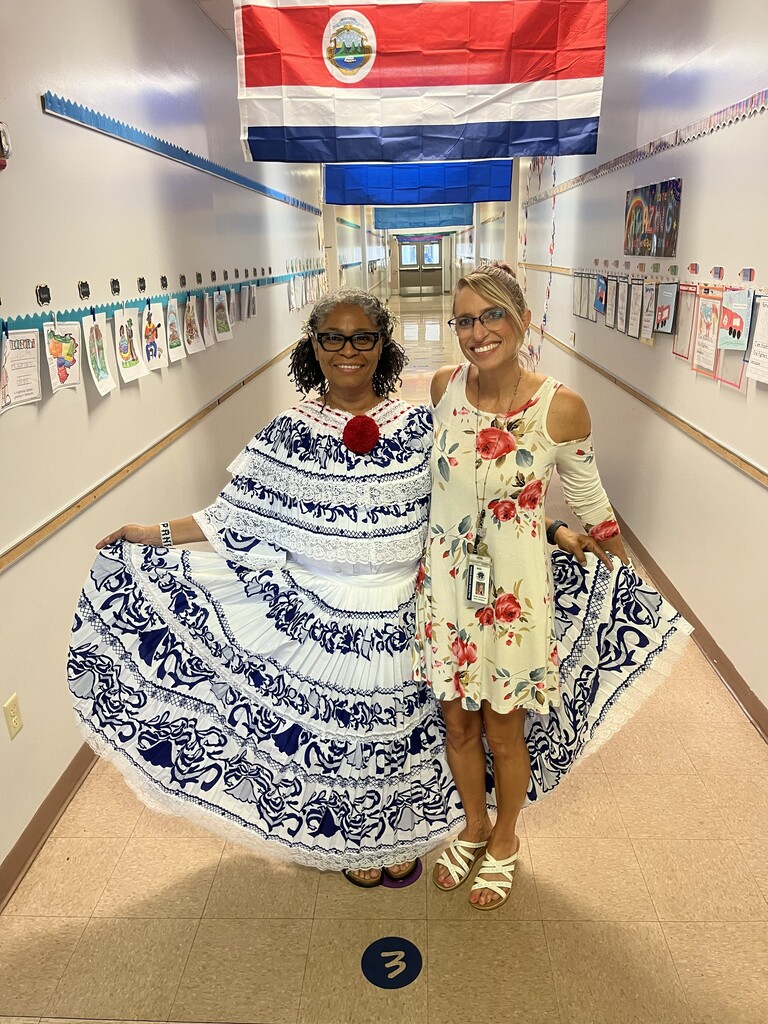 Two women pose in a school hallway; one wears a traditional blue and white dress, the other a floral dress. Flag hangs above them.