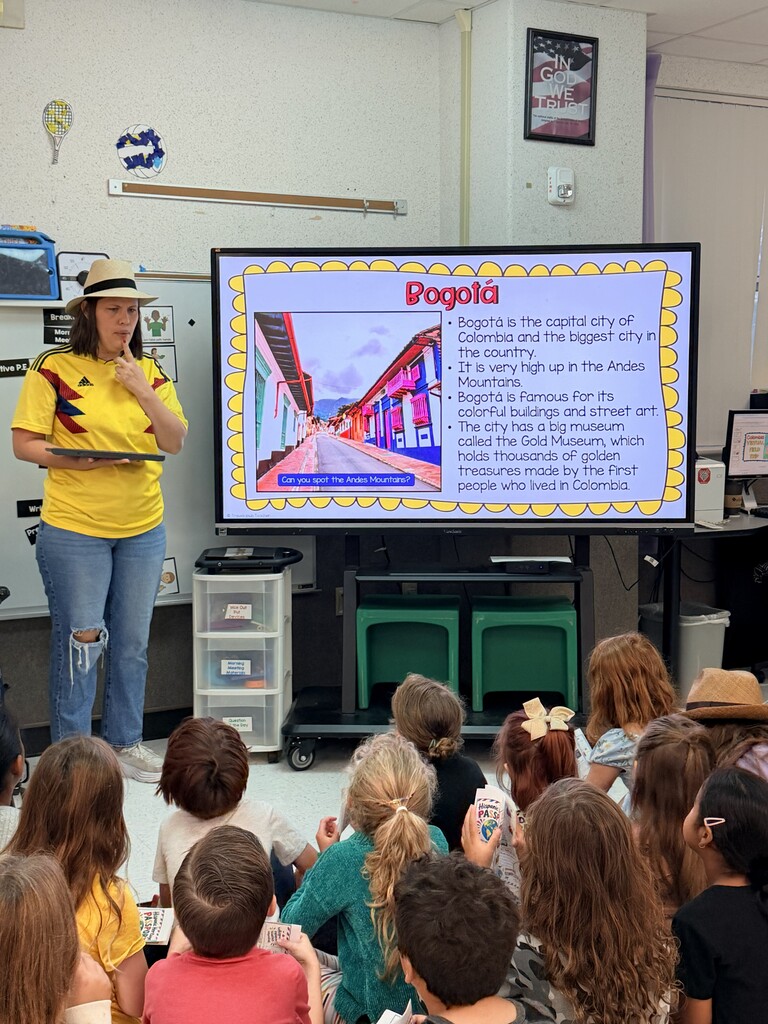 A woman presents to a classroom of children using a projector screen, showing a slide about Bogotá.